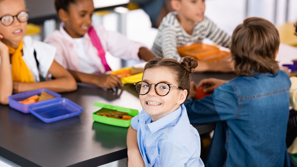 selective focus of excited schoolgirl in eyeglasses looking at camera in school canteen near multiethnic classmates