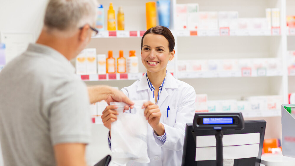 apothecary selling drug to senior man at pharmacy medicine, pharmaceutics, health care and people concept - female apothecary giving senior male customer his purchase in bag at drugstore