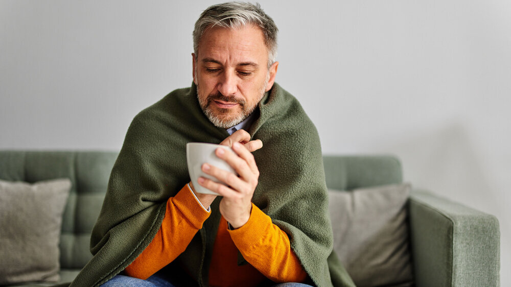 A sick bearded man wrapped in a blanket having a cup of warm tea at home. An unshaven senior man sitting on the couch at home, feeling sick, having a fever, holding a cup of tea.