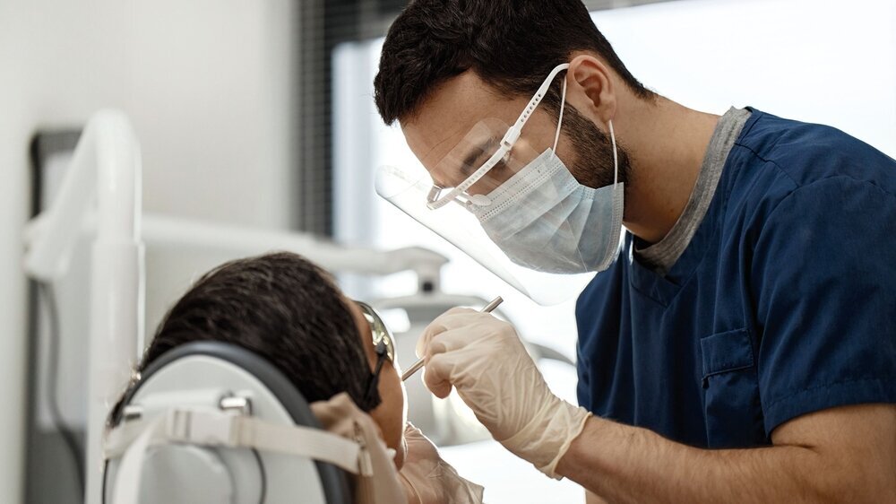 Young Adult Male Dentist Examining Patient during Dental Appointment Young adult male dentist wearing protective mask and face shield treating patient in dental office, using dental instruments while focused on dental procedure, patient reclining in dental chair