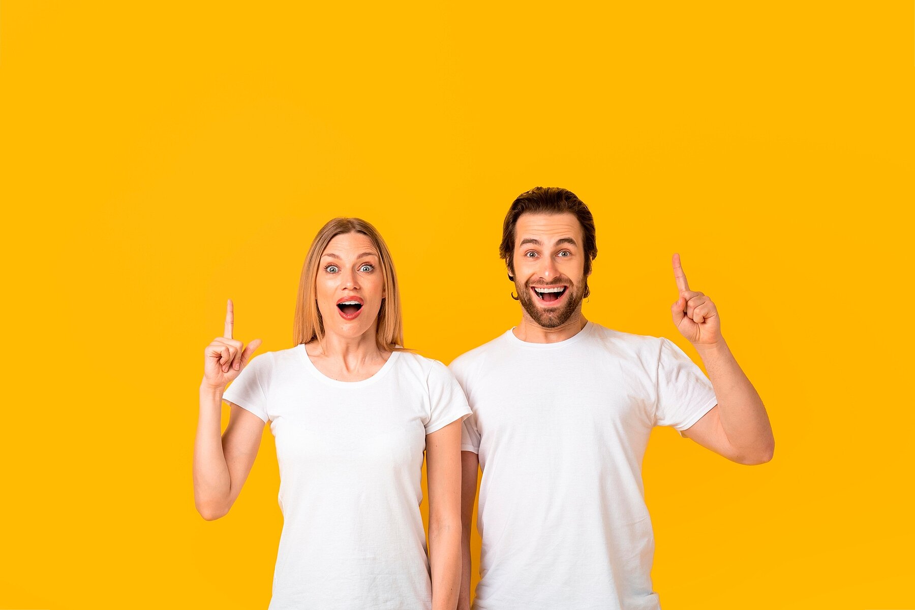 Excited Happy Mixed Gender Couple Smiling And Pointing Upward At Yellow Background, Engaging In Playful Communication, Capturing Joyful Emotion Expressing Enthusiasm In Studio Setting.