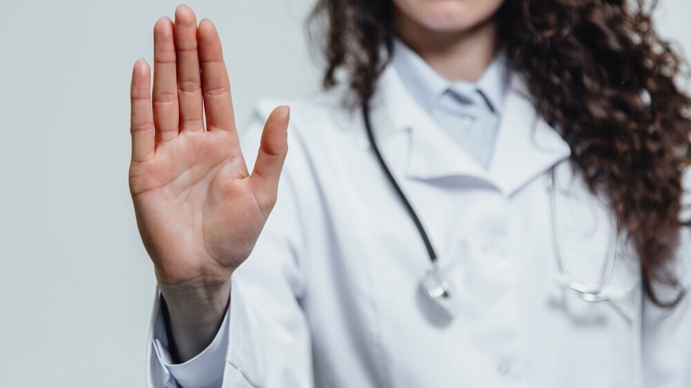 Junge Ärztin hält schützend die Hand erhoben, um Gewalt seitens Patienten abzuwehren.