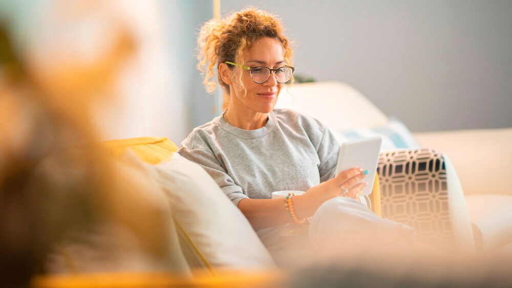 Junge Frau lächelt und genießt es, zu Hause auf dem Sofa sitzend gemütlich ein E-Book zu lesen. Frauen entspannen sich zu Hause.