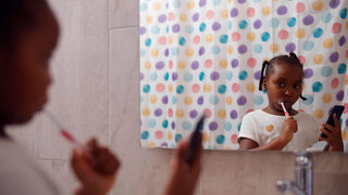 Kleines Mädchen mit Migrationshintergrund putzt sich die Zähne mit Handy in der Hand.