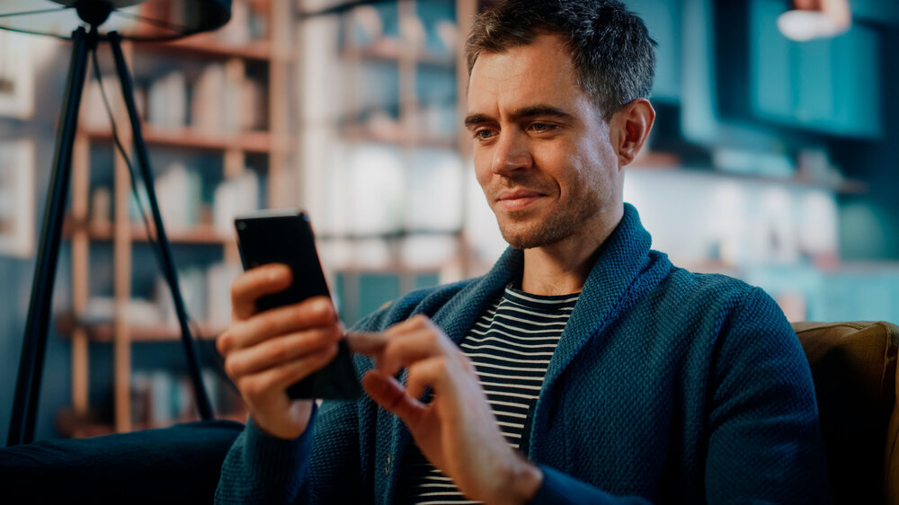 Excited Caucasian Man Using Smartphone while Resting on a Sofa in Stylish Living Room. Happy Man Smiling at Home and Chatting to Colleagues and Clients Over the Internet. Using Social Networks. Mann hat in seiner Freizeit sein Handy in der Hand.