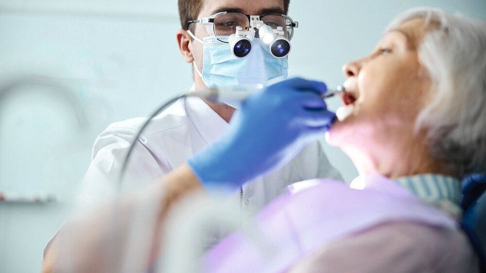 Dentist in dental headlights loupes treating old woman teeth Dental professional attending to elderly citizen mouth cavity with a steel bur on handpiece working on teeth