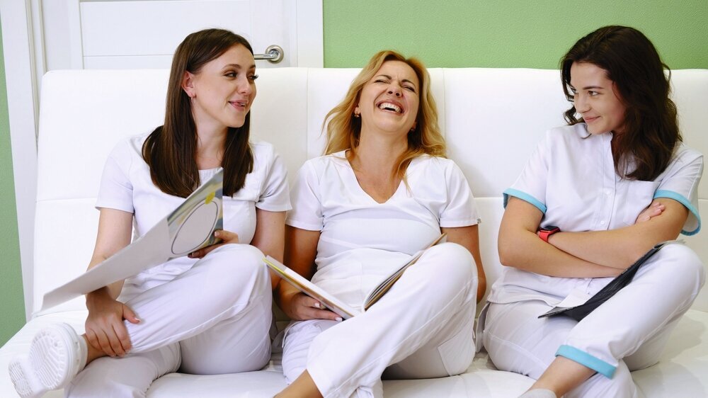 Gesundheitspersonal in weißen Uniformen sitzt während einer Pause zusammen auf der Couch und lacht.