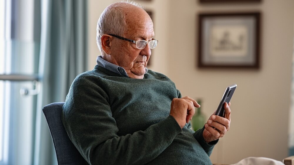 Old man wearing eyeglasses to use smartphone at home. Handsome senior man at home relaxing on couch reading a message on cellphone. Senior sitting alone in his living room and using mobile phone.