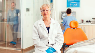 Ältere Zahnärztin sitzt im Behandlungsraum mit Schutzbrille und Kittel. Im Hintergrund läuft der Praxisbetrieb.