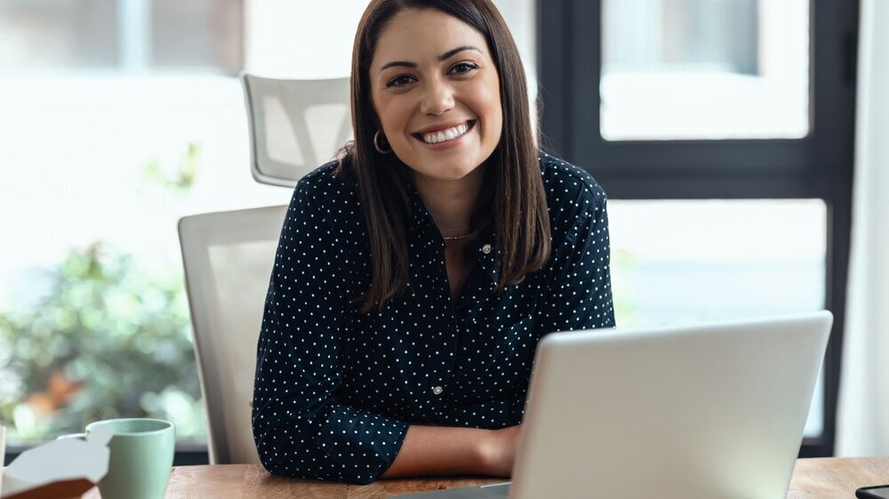 1348209421 Shot of smiling business woman working with laptop while looking at camera in modern startup office.