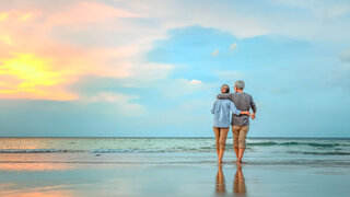 Ein Pärchen spaziert am Strand: Eine ältere Frau und ein älterer Mann mit grauen Haaren schlendern Arm in Arm am Meer. Im Hintergrund ist der Sonnenuntergang.