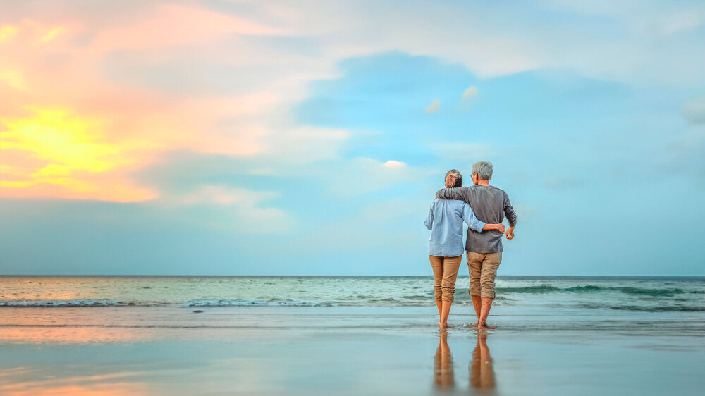Plan life insurance of happy retirement concepts. Senior couple walking on the beach holding hands at beach sunrise in evening. Ein Pärchen spaziert am Strand: Eine ältere Frau und ein älterer Mann mit grauen Haaren schlendern Arm in Arm am Meer. Im Hintergrund ist der Sonnenuntergang.