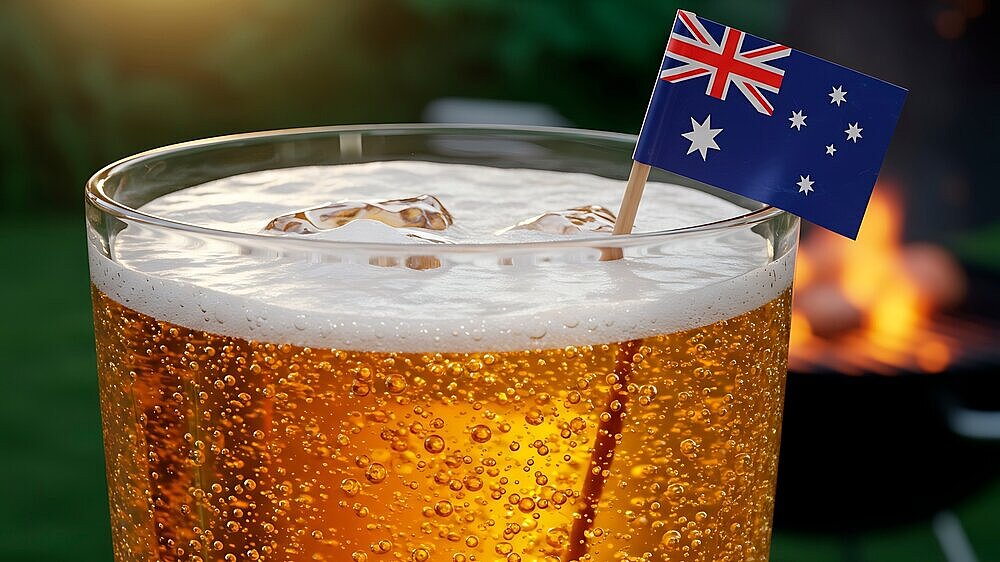 Closeup of a beer glass filled with light golden beer, topped with a frothy head, and ice. A small Australian flag is affixed to a toothpick and in the drink. The background includes an out-of-focus view of a backyard barbecue and green foliage. The warm sunlight highlights the scene. Great for commercial use in advertisements for beverages, outdoor events, or related industries.
