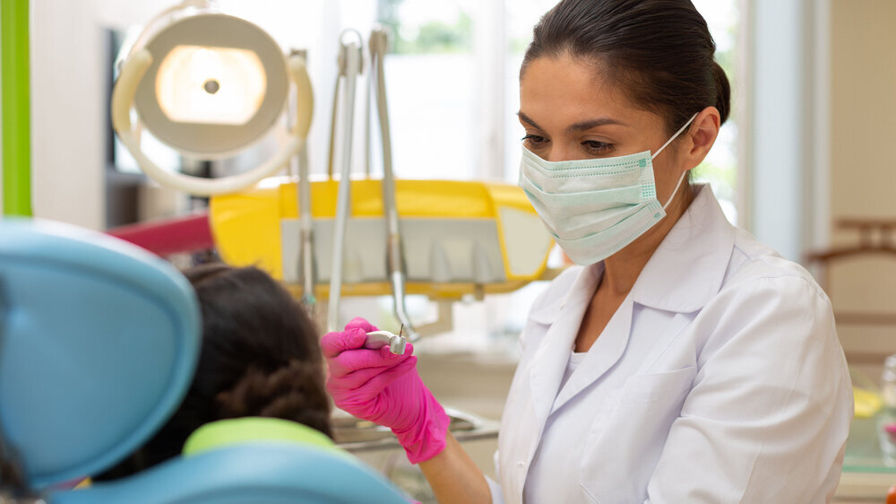 Serious doctor looking at a young female patient Treating caries. Serious woman doctor in pink aseptic gloves looking at a young female patient while holding a dental handpiece