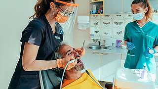 Männlicher Patient, der auf einem Zahnarztstuhl sitzt und von einem Zahnarzt mit Unterstützung einer Dentalhygienikerin eine Zahnsanierung erhält