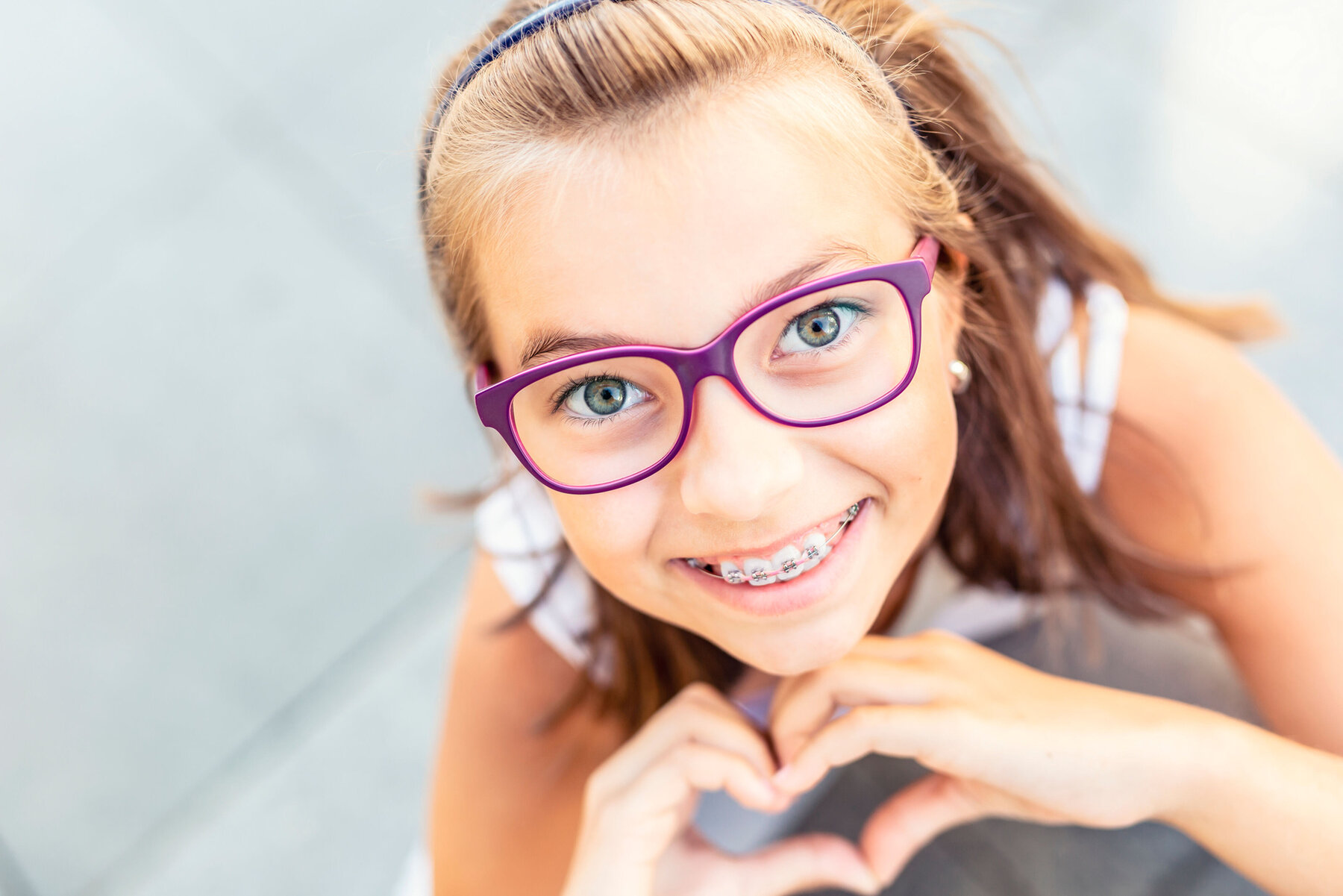 Young preteen girl in glasses wearing braces smiles at the camera showing heart shape with her hands.