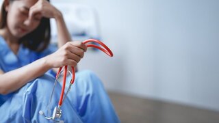 Junge Ärztin in blauem Kittel fühlt sich überwältigt und gestresst, während sie in einem Krankenhauszimmer auf dem Boden sitzt