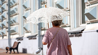 Old woman with sun umbrella walk on a street. Hot weather, life of elderly people in summer