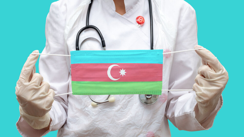 Epidemic in Azerbaijan. Young woman doctor in a medical coat (suit) and gloves holds a medical mask with the print of the flag of Azerbaijan on a blue background isolated. Arzt aus Aserbaidschan