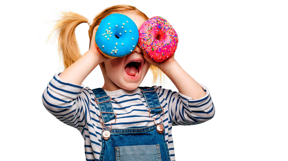 Kleines, blondes Mädchen hält sich zwei bunte Doudhnuts vor die Augen und lächelt. Kleines, blondes Mädchen hält sich zwei bunte Doudhnuts vor die Augen und lächelt.