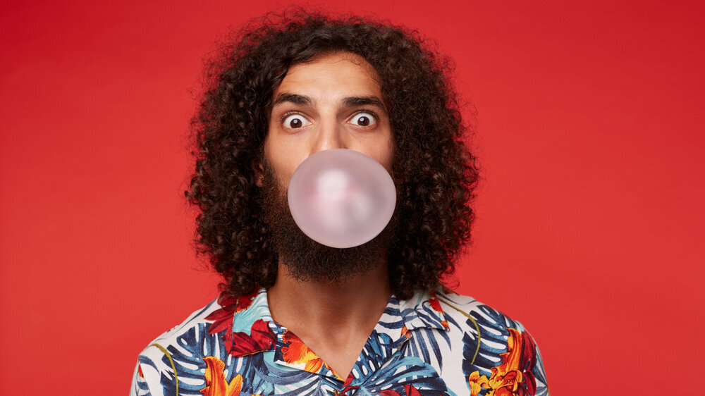 Astonished attractive young brunette curly man with beard wearing multi-colored flowered shirt while posing over red background, rounding eyes and inflating bubble with pink gum