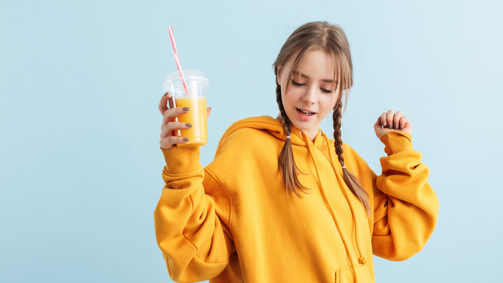 Young attractive girl with two braids in orange hoodie holding plastic cup with juice in hand dreamily dancing over blue background