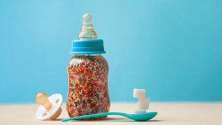 Unhealty baby food concept. The problem of hidden sugar in baby food and drinks. Baby bottle with multicolored sugar dragee on blue background with blank space for text.