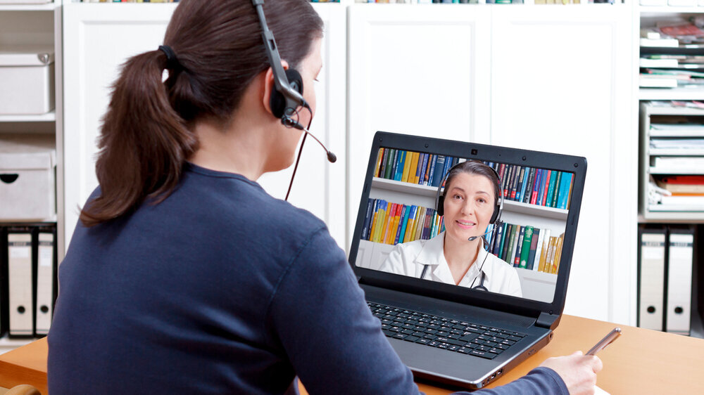 Woman with pen and paper and a headset in front of her computer making a video call with her doctor, e-health, telehealth or telemedicine concept