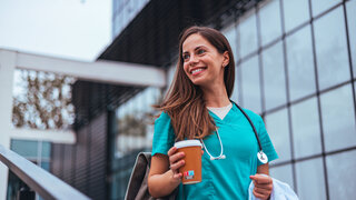Eine lächelnde Krankenschwester in Kittel und mit Stethoskop genießt eine Pause mit einer Kaffeetasse in der Hand vor einer medizinischen Einrichtung.