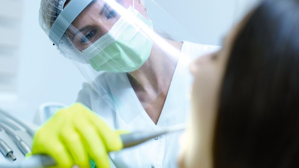 Female dentist with face mask and visor as coronavirus safety precaution checkup dental health of young woman Zahnärztin PSA