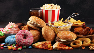 Hochverarbeitete Lebensmittel wie Burger, Doughnuts und Cup Cakes sind bunt aufgetürmt vor schwarzem Hintergrund.