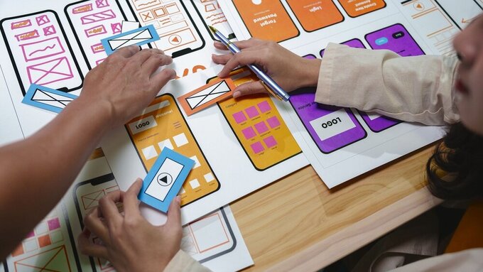 Web designers arranging colorful paper cutouts with application icons and logos on a large sheet of paper while planning user interface and experience for mobile app development Webdesign