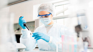 Serious concentrated female microbiologist in sterile clothing and safety goggles sitting at table and dropping reagent in petri dish while doing research in laboratory