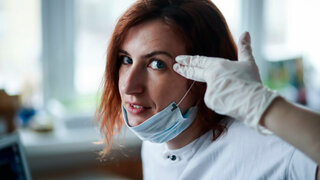 Frau mit Einmalhandschuhen und OP-Maske macht Kopfschussgeste