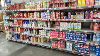 supermarket shelf with a large selection of drinks and dairy products in tetra pack cardboard packaging. The shelves feature different brands and types of products: milkshakes, yoghurts, cream, milk, as well as plant-based alternatives — soy, oat, almond milk. The packaging is brightly colored: pink, yellow, white, blue, and green, creating a rich palette. Popular brands such as Domik v Derevne, Selo Zelenoe, Valio, Bite, Nemoloko, and others are visible. The bottom row also features yoghurts and desserts in plastic cups. The atmosphere of the photo emphasizes the variety of products and the modern retail format.