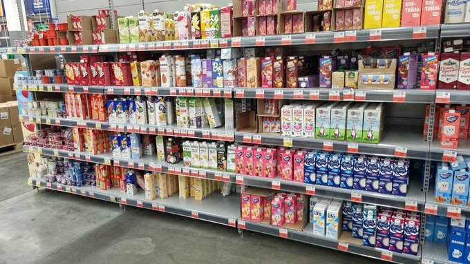 supermarket shelf with a large selection of drinks and dairy products in tetra pack cardboard packaging. The shelves feature different brands and types of products: milkshakes, yoghurts, cream, milk, as well as plant-based alternatives — soy, oat, almond milk. The packaging is brightly colored: pink, yellow, white, blue, and green, creating a rich palette. Popular brands such as Domik v Derevne, Selo Zelenoe, Valio, Bite, Nemoloko, and others are visible. The bottom row also features yoghurts and desserts in plastic cups. The atmosphere of the photo emphasizes the variety of products and the modern retail format.