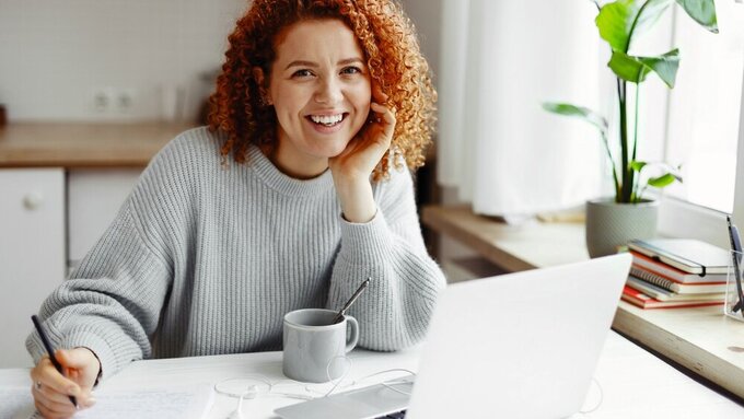 Portrait of smiling redhead curly female student studying at home, doing home work in front of laptop, preparing for exams, sitting at kitchen table with pen in hands, looking at camera