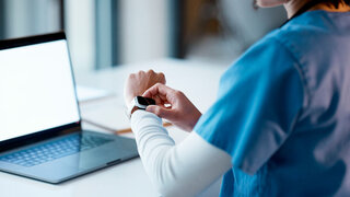Frau im Arztkittel vor ihrem Laptop schaut auf ihre Smartwatch