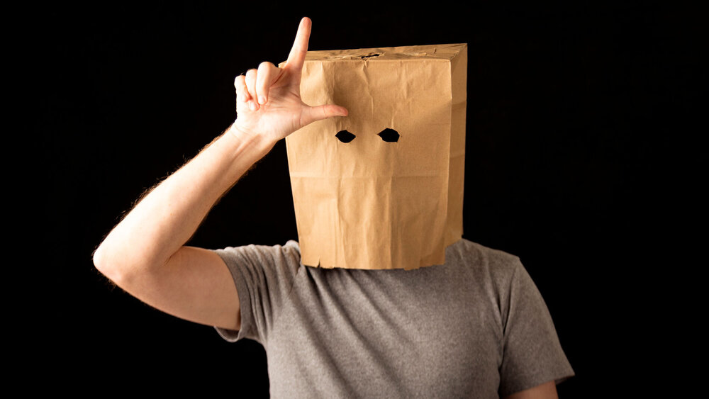 Mann im T-Shirt trägt braune Papiertüte auf dem Kopf und macht das Losersymbol.