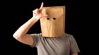 Mann im T-Shirt trägt braune Papiertüte auf dem Kopf und macht das Losersymbol.