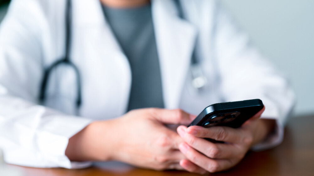 Close-up view of doctor using smartphone to access electronic health records, manage appointments, and communicate with patients, highlighting mobile integration in the healthcare system. Arzt mit Smartphone
