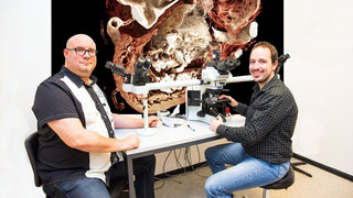 Die Fotomontage zeigt PD Dr. Mark Kühnel (links) und Christopher Werlein an einem Mehrender-Mikroskop mit einem Gewebeschnitt der durch COVID-19 umgebauten Herzgefäße.