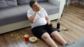 Übergewichtiger Junge sitzt essend vor dem Fernseher ans Sofo gelehnt.