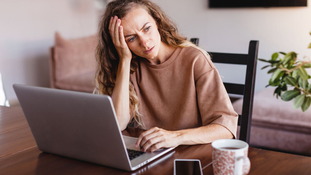 Junge Frau sitzt vor dem Laptop und fühlt sich krank, wirkt abgeschlagen und gestresst.