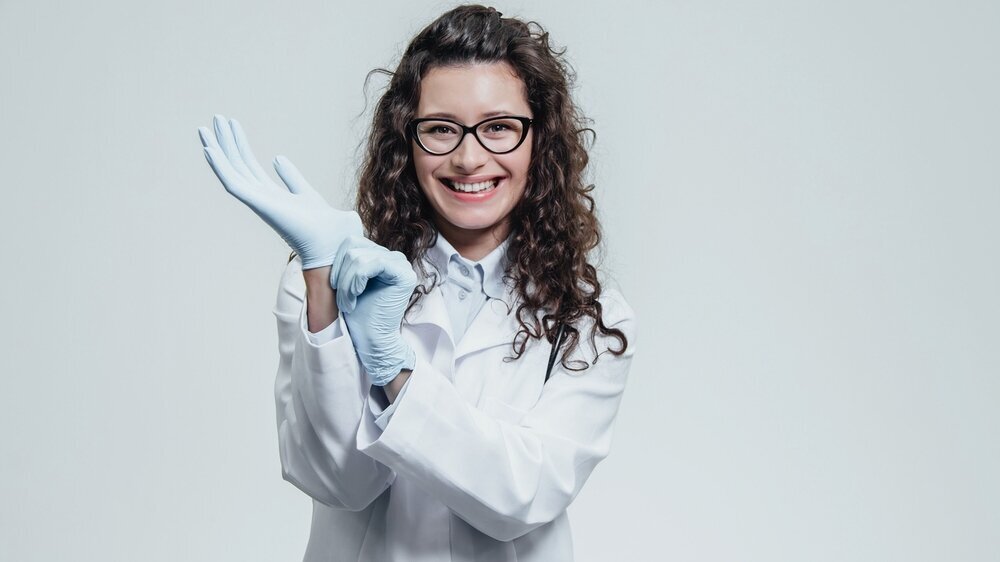 Eine junge Ärztin mit Brille und blauen Handschuhen blickt aufmerksam in die Kamera. Eine junge Ärztin mit Brille und blauen Handschuhen blickt aufmerksam in die Kamera.