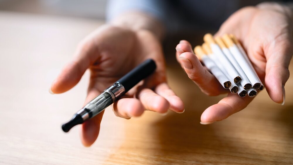 Die Hand einer Frau hält Vaporizer und Tabakzigaretten über hölzernem Schreibtisch Bisherige Studien zum Nutzen von E-Zigaretten bei der Rauchentwöhnung brachten bislang unterschiedliche Ergebnisse. Eine aktuelle US-Analyse spricht ihnen einen Nutzen eher ab.