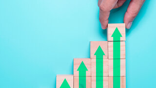 Themenbild Kostensteigerung: Hand legt Holzwürfel aufeinander, die einen größer werdenden Pfeil nach oben ergeben