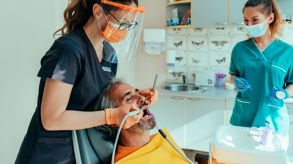 Männlicher Patient, der auf einem Zahnarztstuhl sitzt und von einem Zahnarzt mit Unterstützung einer Dentalhygienikerin eine Zahnsanierung erhält Männlicher Patient, der auf einem Zahnarztstuhl sitzt und von einem Zahnarzt mit Unterstützung einer Dentalhygienikerin eine Zahnsanierung erhält