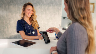 Smiling doctor accepting contactless payment from female patient at modern clinic reception desk, using nfc technology for quick and easy transaction