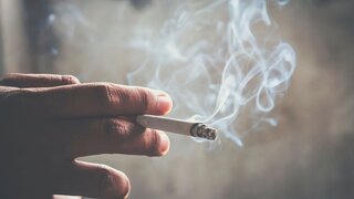 Mann hält qualmende Zigarette in der Hand. Rauch steigt auf.